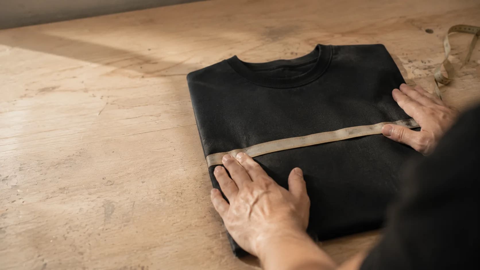 A tape measure laid across the chest of a folded dark garment while two hands smooth the fabric flat