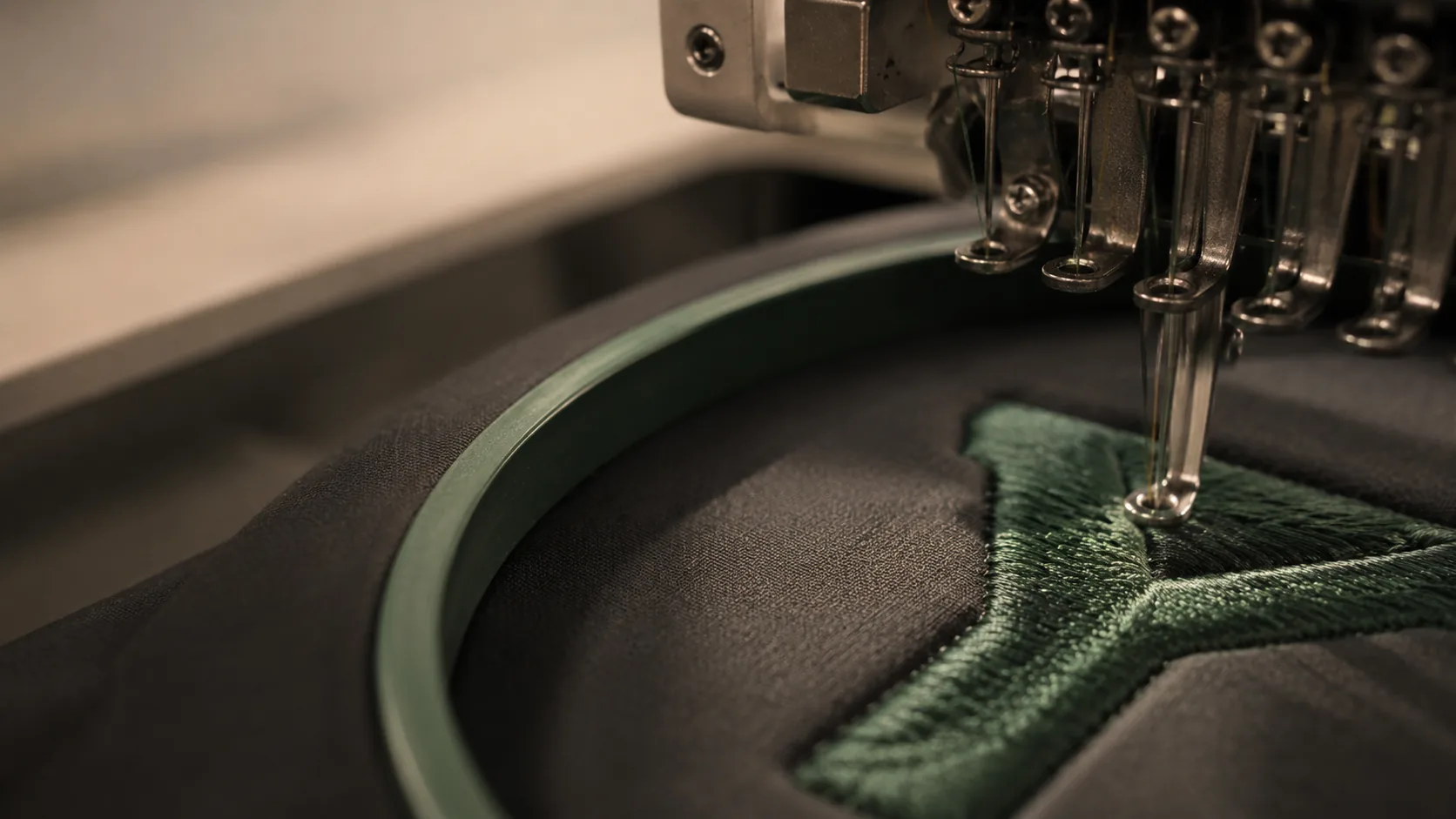 A close macro of dense satin-stitch embroidery being formed in green thread on a dark garment held in the hoop, with the needle bar directly above it